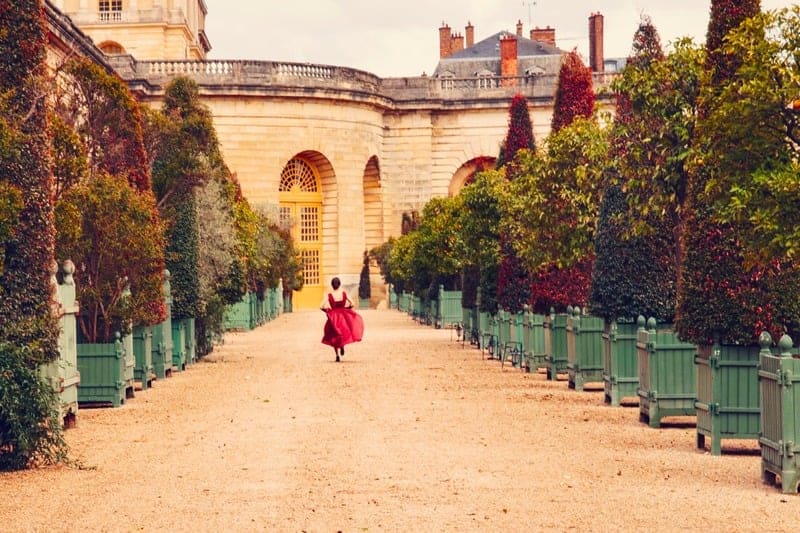 Invitée en robe rouge dans une allée bordée d’arbustes du Château de Versailles